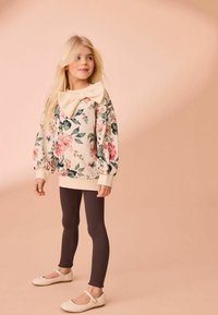 Krem sweatshirt s cvetnim vzorcem in velikim voščilom na ovratniku, v kombinaciji z temnimi rebrastimi leginami in kremnimi Mary Jane čevlji. Gladka tekstura.
