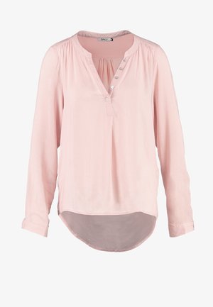 Helle rosa Bluse mit V-Ausschnitt und Knopfdetails, kombiniert mit dunklen Jeans und tanfarbenen Ankle Boots, hält einen braunen Hut. Glatte Textur.