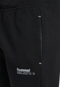 Czarne spodnie sportowe z detalami przy bocznym szwie oraz białym napisem z logo "hummel" w pobliżu krawędzi kieszeni na górnej części uda.