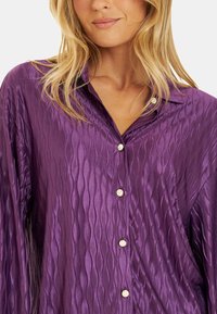 Blusa de satén morado con un patrón de olas texturizado, cuello clásico y botones dorados en la parte delantera y la tapeta.