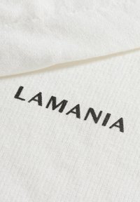 Fehér textil, amelyen a "LAMANIA" szó vastag fekete hímzéssel szerepel, sima, enyhén struktúrált felülettel és finom szövésmintával.