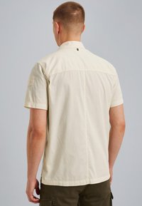 Chemise à manches courtes, beige clair, avec un col standard, texture unie et un petit logo dans le dos, associée à un pantalon cargo.