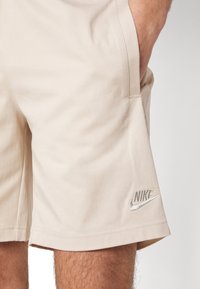 Beige Nike shorts made from cotton blend material, featuring a silver embroidered logo on the left leg and side pockets.