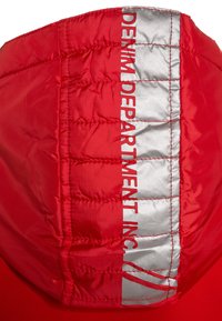 Capuche rembourrée rouge avec surface texturée, comportant une bande verticale argentée avec "DENIM DEPARTMENT INC." imprimé en rouge.