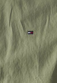 Olivengroene stof met een klein, geborduurd logo en blauwe, witte en rode strepen. De textuur lijkt licht gekreukt.