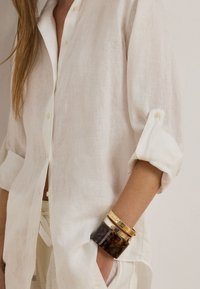 Linen button-up shirt in off-white, rolled sleeves, featuring light texture. Accompanied by stacked gold and tortoiseshell bangles.