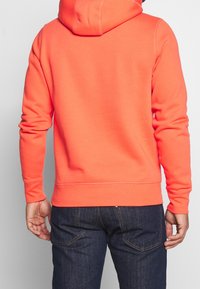 Felpa arancione brillante con cappuccio regolabile e polsini a coste, mostrata di spalle, abbinata a jeans blu scuro. Texture morbida e design semplice.