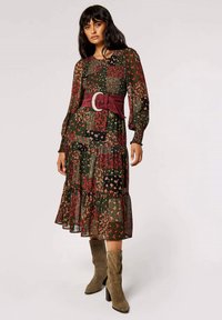 Ein Blumenmuster-Kleid mit langen Ärmeln, grünem Grundton, roten und gelben Blumen, gestuftem Saum und einem breiten bordeauxfarbenen Gürtel. Kombiniert mit olivgrünen Knöchelstiefeln.