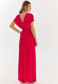 Kobieta z brązowymi włosami w niskim kucyku, ubrana w jasnoróżową, długa suknię z głębokim dekoltem z tyłu i szerokim pasem w talii, stojąca na tle białym.