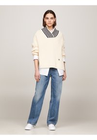 Pull en tricot couleur crème avec un décolleté plongeant et des rayures bleu marine, assorti à un jean large bleu clair et des baskets blanches.