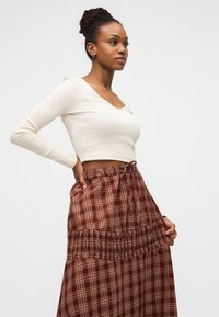 Mlada žena s naušnicama koja nosi kremasti uski duži top s dugim rukavima i smeđu kariranu suknju visokog struka, pozira s rukom na bokovima ispred bijele pozadine.