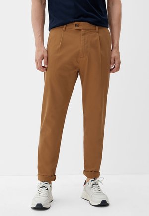 Man draagt opgerolde bruine broek, witte sneakers en een ingestopt donkerblauw T-shirt, staand tegen een effen witte achtergrond.