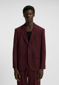 Burgundy blazer made of smooth fabric with a single button, notched lapels, and side pockets, styled over a black top.