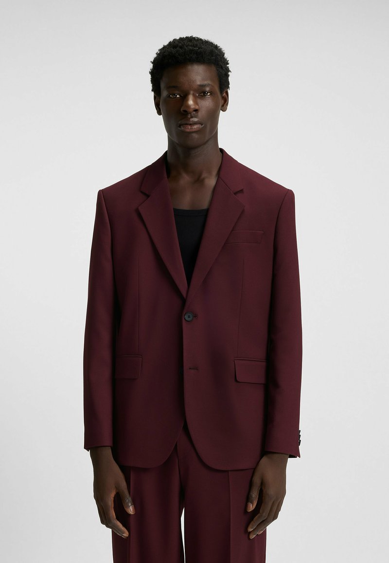 Burgundy blazer made of smooth fabric with a single button, notched lapels, and side pockets, styled over a black top.