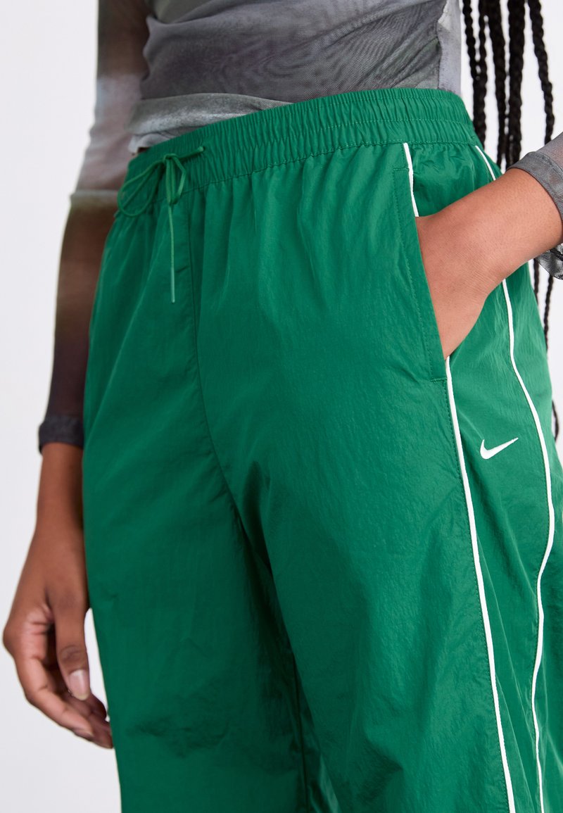 Groene sportbroek van lichtgewicht, gestructureerde stof, met een elastische tailleband, zijzakken en witte piping langs de naden. Nike-logo aanwezig.