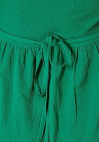 Groene stoffen jurk met een textuurafwerking, een elastische tailleband en een knoopriemaccent. Het ontwerp heeft vloeiende plooien bij de taille.
