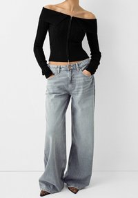 Kvinna i svart stickad zip-up topp med bar axel, ljusa vida jeans och spetsskor med leopardmönster, händerna i fickorna.