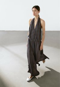 Black and blue plaid halter jumpsuit with a deep V-neck, fitted waist, and loose, flowing legs. White flat shoes. Plain background.