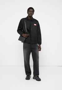 Svart jakke med rødt "DIESEL"-logo, sammen med mørkegrå jeans og svarte sneakers. Vesken er også svart med preget "DIESEL"-merke.