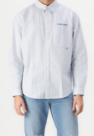 Muž nosící světle modrou a bíle pruhovanou košili s knoflíky a logem "Tommy Jeans" na kapsičce na hrudi, v kombinaci se světle modrými roztrhanými džíny.