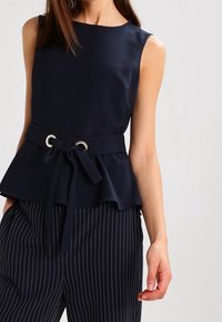 Topp i marineblått med peplum og rund hals, stoffbelte i midjen, og sølvmetallakenter. Kombinert med bukser med smale striper.