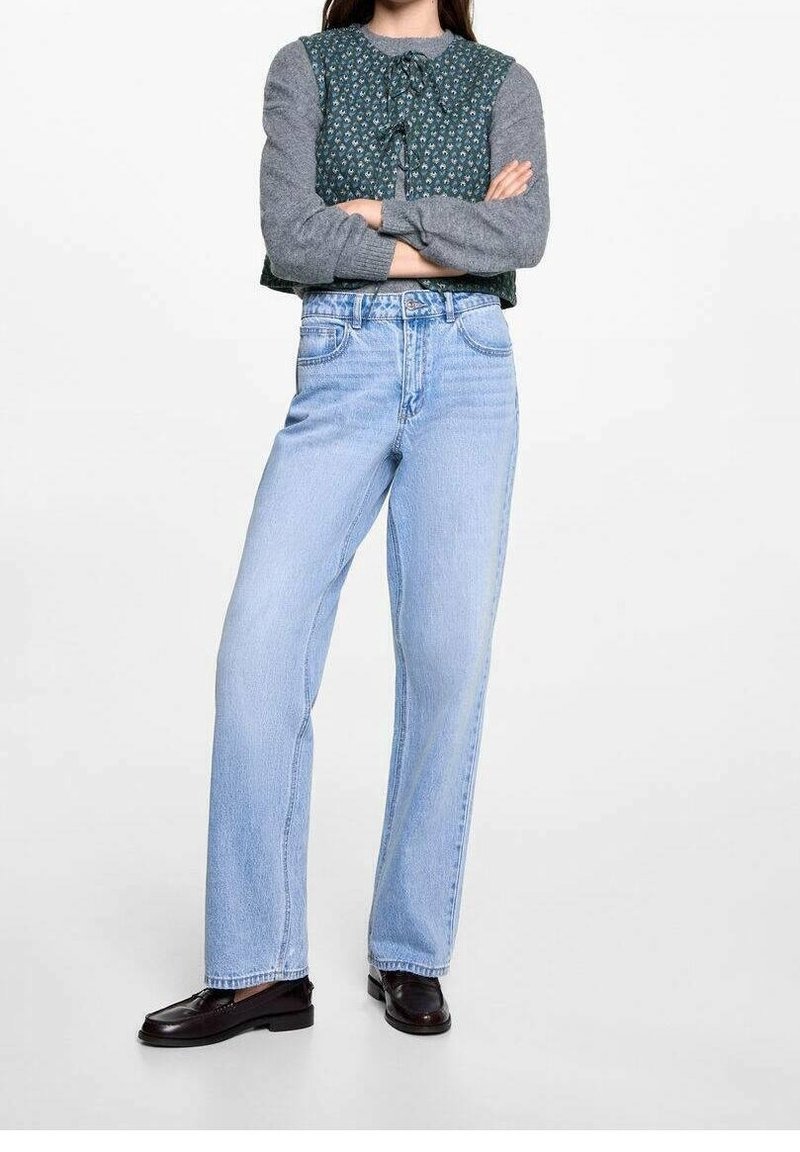 Jasnoniebieskie jeansy o szerokich nogawkach zestawione z szarym długim topem, który ma zieloną wzorzystą kamizelkę na wierzchu oraz kokardkę z przodu.