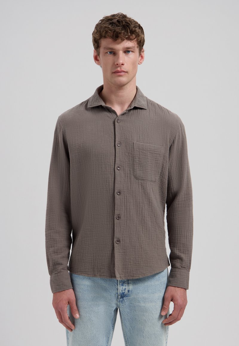 Jeune homme aux cheveux bouclés portant une chemise marron texturée à manches longues avec boutons et un jean bleu clair, posé devant un fond uni.