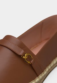 Mocassin en cuir marron avec un détail en anneaux entrelacés dorés et une semelle en jute tressé, présenté en gros plan.