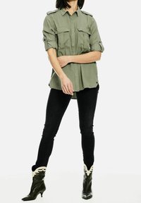 Camicia verde oliva con bottoni, dotata di due tasche sul petto, maniche arrotolate e una vestibilità rilassata, abbinata a jeans skinny neri e stivaletti neri con punta appuntita.