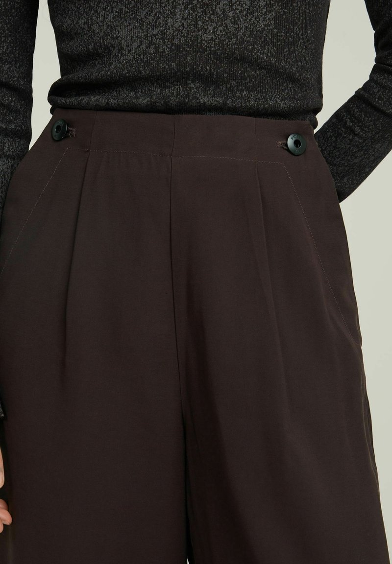 Brązowe spodnie z zakładkami z dwoma czarnymi guzikami w pasie oraz kieszeniami bocznymi. Materiał jest gładki z delikatnym połyskiem.