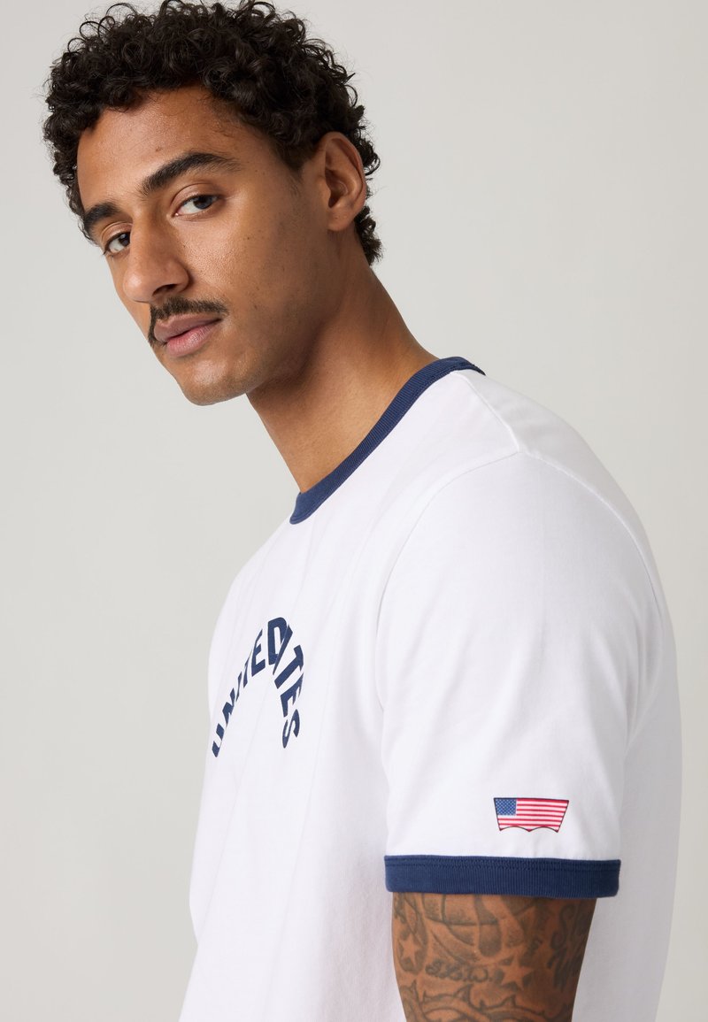 Man with curly hair and mustache wearing white T-shirt with navy trim, "United Ties" text, and small American flag patch on sleeve.