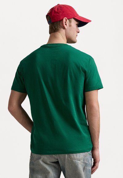 Πράσινο βαμβακερό t-shirt με στρογγυλή λαιμόκοψη, κοντά μανίκια και χαλαρή γραμμή, συνδυασμένο με μια κόκκινη καπέλο και ανοιχτό μπλε τζιν παντελόνι.