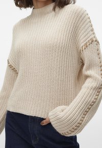 Beżowy sweter dzianinowy z wysokim dekoltem i prążkowanymi rękawami, z dekoracyjnym otwartym splotem wzdłuż szwów ramion oraz luźnym krojem.