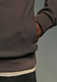 Brun quiltet sweatshirt med en tekstureret overflade, ribbede manchetter og en lagdelt beige undertrøje synlig ved sømmen.