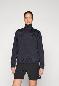 Donkerblauwe windbreakerjas met een hoge kraag, half-rits sluiting, elastische manchetten en een cinchzoom. Gecombineerd met zwarte sportshorts.
