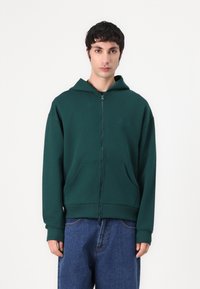 Grønn zip-up hettegenser laget av mykt stoff, med kengurulomme, ribbede mansjetter og hette. Pr präget logo detalj på brystet.