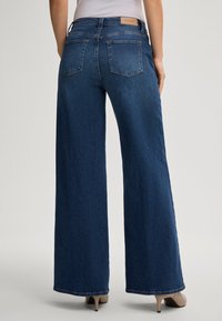 Jeans in denim scuro a gamba ampia con vita alta, dotati di due tasche posteriori e di una patch in pelle sul cintura. Tessuto morbido.