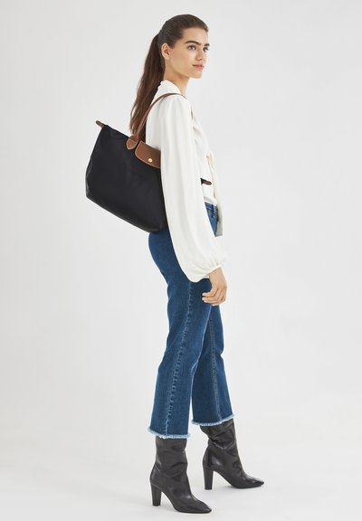 Black tote bag with brown leather accents, featuring a smooth fabric exterior and a spacious design, held by a shoulder strap.