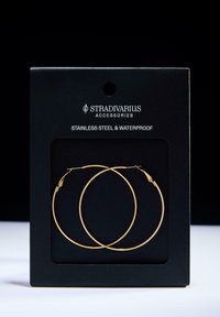 Zlati uhani iz nerjavečega jekla v obliki obroča, vodoodporni, predstavljeni na črni kartici z blagovno znamko "Stradivarius Accessories". Preprost in krožen dizajn.