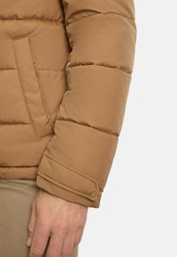 Tannfarbener gepolsterter Jackenärmel mit glatter Textur, der ein gesticktes Design und eine Seitenasche aufweist. Der Ärmel hat einen umgebogenen Rand.