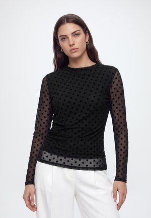 Femme portant un haut noir transparent à pois à manches longues et un pantalon blanc, se tenant devant un fond clair uni.