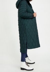 Abrigo largo verde acolchado con capucha, botones de presión en la parte delantera y aberturas laterales. Llevado sobre vaqueros azules y botas blancas de cordones hasta el tobillo.