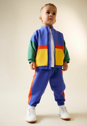 Mehrfarbiger Trainingsanzug mit einer blauen Jacke mit Reißverschluss, grünen Ärmeln, gelben und roten Akzenten sowie passenden blauen Hosen mit orangen Streifen.