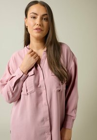 Camisa abotonada de color rosa claro con mangas largas, dos bolsillos frontales y un corte relajado. La tela parece suave y ligeramente texturizada.