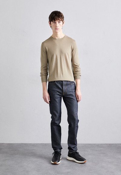 Camisa de manga larga marrón claro combinada con jeans azul oscuro. El modelo está de pie en zapatillas negras sobre un fondo liso y texturizado.