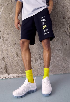 Henkilö, joka pukeutuu mustiin Nike-logoshortseihin, kirkkaankeltaisiin sukkiin ja valkoisiin Nike-tennareihin, seisoo teksturoitua betoniseinää vasten.
