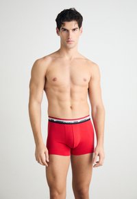 Rote Baumwoll-Boxershorts mit einem schwarz-weißen Markengummibund, die eine enge Passform und eine glatte Textur aufweisen, getragen von einem Modell.