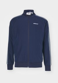Tengerészeti kék, cipzáras sportdzseki sima anyagból, fehér háromsávos díszítéssel az ujjakon és hímzett logóval a mellrészen.