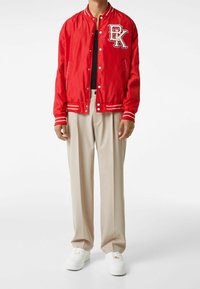 Rode bomberjack met witte gestreepte manchetten, voorzien van een geborduurd logo. Gekozen boven een zwart topje, gecombineerd met beige broeken en witte sneakers.
