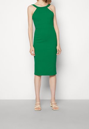 Femme portant une robe verte sans manches, longueur genou, avec des découpes aux épaules, et des sandales beiges à talons blocs avec brides.